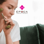 Uma mulher está usando um massageador facial enquanto está sentada em um ambiente doméstico. No topo da imagem, estão as logos da Livelo e da Época Cosméticos.