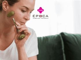 Uma mulher está usando um massageador facial enquanto está sentada em um ambiente doméstico. No topo da imagem, estão as logos da Livelo e da Época Cosméticos.
