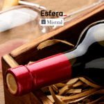 Uma garrafa de vinho com selo vermelho está deitada em uma caixa de madeira, cercada por palha. Ao fundo, há uma taça de vinho inclinada. Na parte superior da imagem estão as logos da Esfera e Mistral.