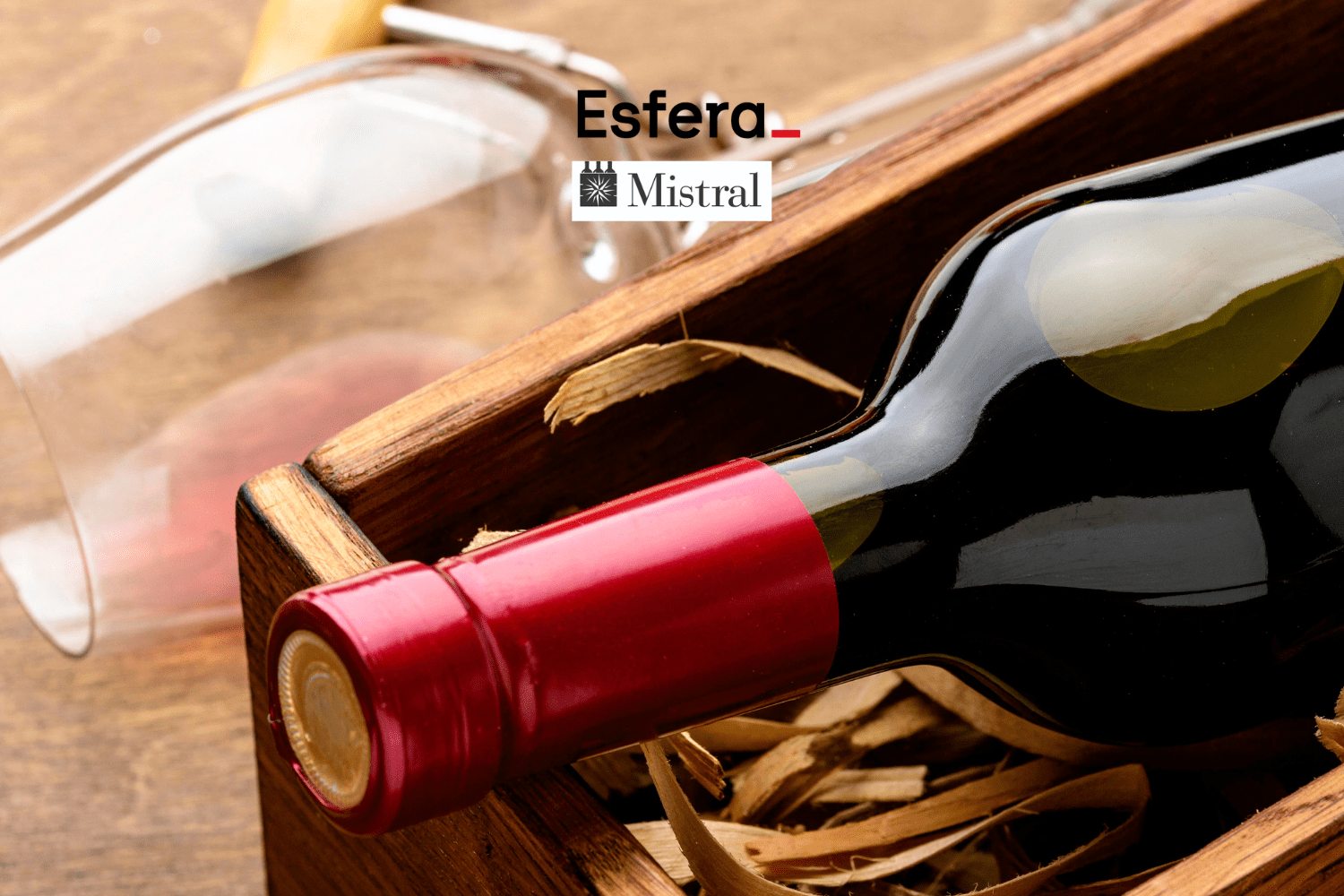 Uma garrafa de vinho com selo vermelho está deitada em uma caixa de madeira, cercada por palha. Ao fundo, há uma taça de vinho inclinada. Na parte superior da imagem estão as logos da Esfera e Mistral.