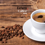 Uma xícara de café espresso sobre um pires branco está posicionada à direita da imagem, sobre uma superfície de madeira. Ao lado, alguns grãos de café estão espalhados. No canto superior esquerdo, estão as logos da Esfera e Coffee++
