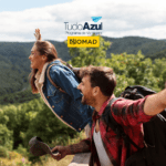 Um casal jovem está sorrindo e com os braços abertos, em uma área montanhosa e arborizada, transmitindo sensação de liberdade. Ambos estão com mochilas, sugerindo uma viagem de aventura. No topo da imagem, estão as logos do "Tudo Azul Programa de Vantagens" e "Nomad".