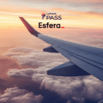 Vista de uma asa de avião voando acima das nuvens durante o pôr do sol. As cores do céu são suaves, variando entre tons de laranja e rosa. No topo da imagem, estão as logos do "LATAM Pass" e "Esfera".