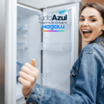 Uma mulher sorridente e animada está de frente para uma geladeira aberta, fazendo um gesto de positivo com o polegar. Ela usa uma jaqueta jeans. No topo da imagem, estão as logos "TudoAzul Programa de Vantagens" e "Magalu".