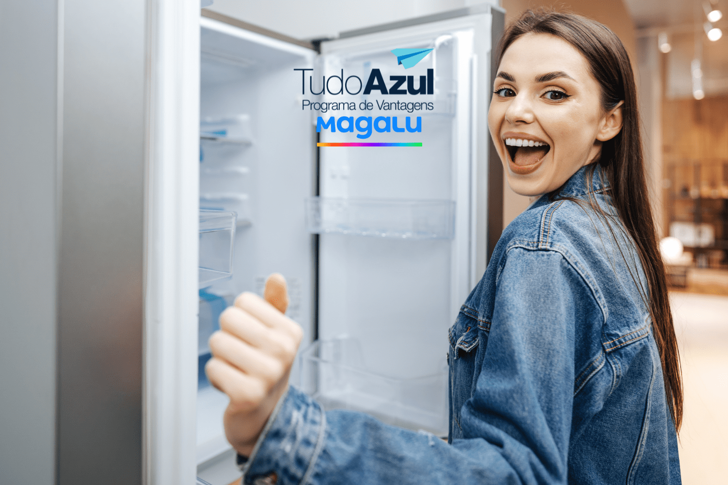 Uma mulher sorridente e animada está de frente para uma geladeira aberta, fazendo um gesto de positivo com o polegar. Ela usa uma jaqueta jeans. No topo da imagem, estão as logos "TudoAzul Programa de Vantagens" e "Magalu".