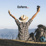 Um viajante de chapéu de palha, sentado de costas, levanta os braços em sinal de comemoração, segurando um binóculo. Ele está em uma paisagem montanhosa, com uma mochila de trilha ao seu lado. No topo da imagem, está a logo da "Esfera".