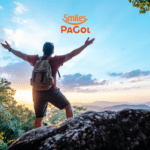 Imagem de um homem com mochila, em pé sobre uma pedra, de braços abertos, apreciando uma vista panorâmica de montanhas ao amanhecer. O céu está claro com algumas nuvens, criando um cenário de natureza exuberante. No topo da imagem, estão as logos da Smiles e PaGol, sugerindo uma parceria entre as duas marcas.