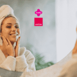 Imagem de uma mulher sorridente, em frente ao espelho, aplicando produtos faciais com as mãos. Ela está vestida com um roupão branco felpudo e uma toalha enrolada na cabeça, transmitindo uma sensação de autocuidado e bem-estar. No centro da imagem estão as logos da Livelo e da Avon.