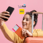 Imagem de uma jovem sorridente tirando uma selfie enquanto segura um passaporte e uma passagem aérea. Ela está ao lado de uma mala de viagem de cor laranja. Ao fundo, a parede rosa suave destaca a cena. No topo da imagem, estão as logos do programa de vantagens TudoAzul e do KM de Vantagens.