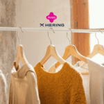 A imagem mostra um cabideiro com várias peças de roupas penduradas, predominando tons neutros e terrosos, com uma peça em destaque na cor mostarda. As roupas são simples e casuais, remetendo a conforto e estilo básico. O ambiente é iluminado pela luz natural que entra pela janela ao fundo, criando uma atmosfera aconchegante. Acima das roupas, aparecem os logotipos da Livelo e da Hering, indicando uma possível colaboração entre as marcas, sugerindo a ideia de acúmulo de pontos ou benefícios ao comprar roupas da Hering através do programa Livelo.