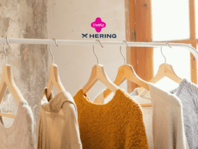 A imagem mostra um cabideiro com várias peças de roupas penduradas, predominando tons neutros e terrosos, com uma peça em destaque na cor mostarda. As roupas são simples e casuais, remetendo a conforto e estilo básico. O ambiente é iluminado pela luz natural que entra pela janela ao fundo, criando uma atmosfera aconchegante. Acima das roupas, aparecem os logotipos da Livelo e da Hering, indicando uma possível colaboração entre as marcas, sugerindo a ideia de acúmulo de pontos ou benefícios ao comprar roupas da Hering através do programa Livelo.