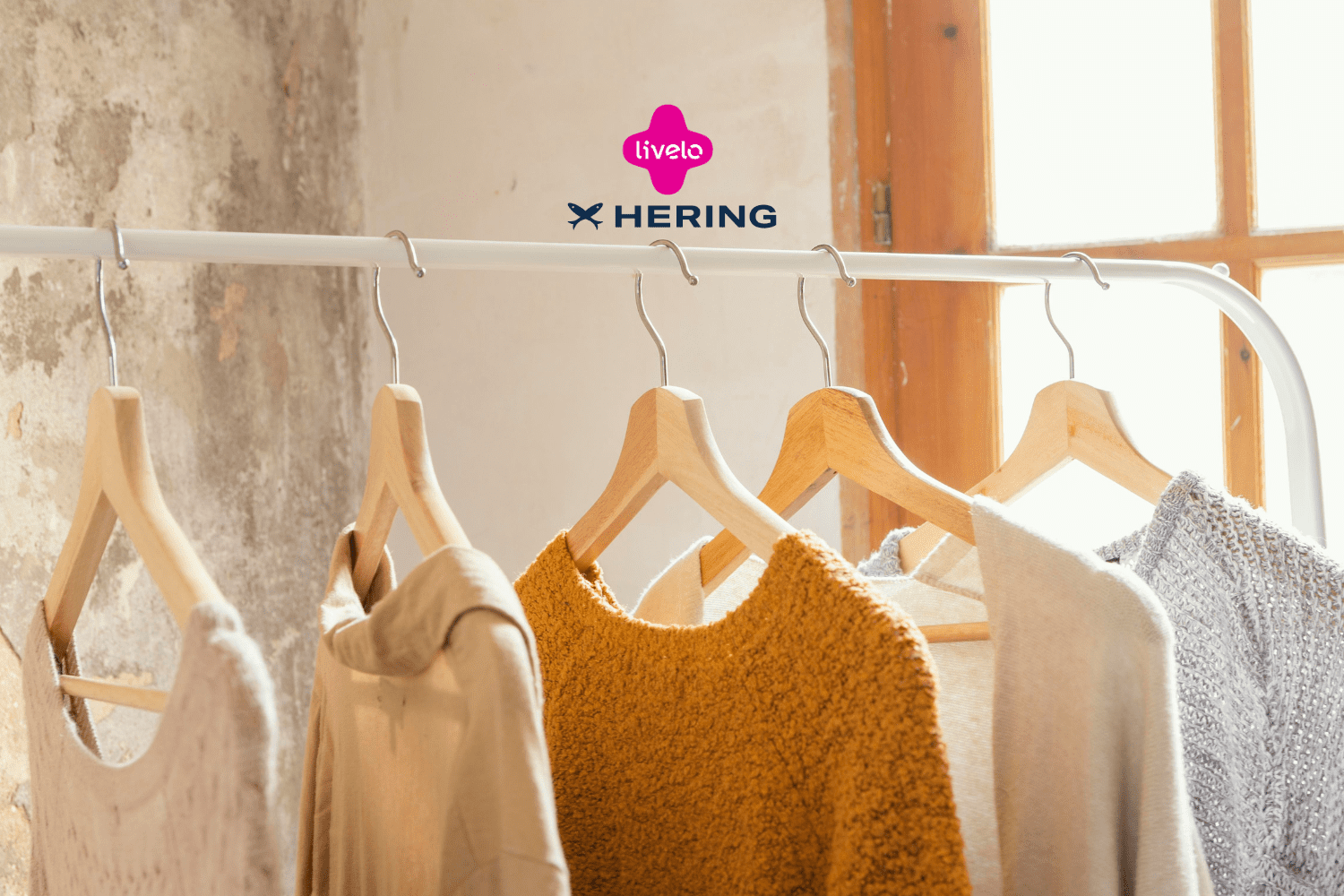 A imagem mostra um cabideiro com várias peças de roupas penduradas, predominando tons neutros e terrosos, com uma peça em destaque na cor mostarda. As roupas são simples e casuais, remetendo a conforto e estilo básico. O ambiente é iluminado pela luz natural que entra pela janela ao fundo, criando uma atmosfera aconchegante. Acima das roupas, aparecem os logotipos da Livelo e da Hering, indicando uma possível colaboração entre as marcas, sugerindo a ideia de acúmulo de pontos ou benefícios ao comprar roupas da Hering através do programa Livelo.