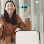 Mulher jovem sorrindo, ajoelhada ao lado de sua mala branca em um aeroporto, segurando um passaporte azul. Ela está vestida com um casaco marrom claro. Ao fundo, um corredor iluminado com grandes janelas. Logotipos da Livelo e CiClic estão inseridos acima, sugerindo uma parceria entre as marcas."