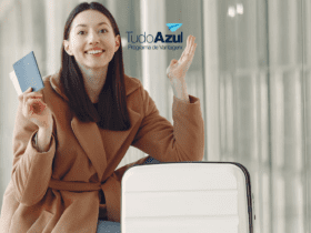Mulher jovem sorrindo, ajoelhada ao lado de sua mala branca em um aeroporto, segurando um passaporte azul. Ela está vestida com um casaco marrom claro. Ao fundo, um corredor iluminado com grandes janelas. Logotipos da Livelo e CiClic estão inseridos acima, sugerindo uma parceria entre as marcas."