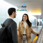 "Em um ambiente de aeroporto, um homem com mochila e uma mulher com um casaco bege estão conversando e sorrindo enquanto caminham em uma esteira rolante. Eles estão em um corredor iluminado por luzes brancas e suaves. Acima deles estão os logotipos do programa de vantagens TudoAzul e da Coopera, sugerindo uma parceria que envolve viagens."
