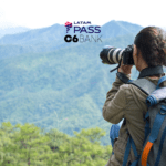 "Uma jovem fotógrafa, equipada com uma mochila e uma câmera profissional, fotografa a paisagem montanhosa à sua frente. O cenário é composto por montanhas verdes sob um céu claro, transmitindo tranquilidade e conexão com a natureza. Acima da imagem, estão os logotipos da LATAM Pass e C6 Bank, sugerindo uma parceria que oferece benefícios para viajantes e entusiastas da fotografia ao ar livre."