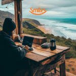 "Uma pessoa trabalha em seu laptop em uma cabana de madeira com vista para uma paisagem costeira deslumbrante, onde o mar azul encontra a praia dourada. O clima parece frio e nublado, com uma garrafa térmica sobre a mesa, criando uma atmosfera aconchegante. Acima, o logotipo da Smiles sugere uma conexão com viagens e experiências ao ar livre."