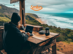 "Uma pessoa trabalha em seu laptop em uma cabana de madeira com vista para uma paisagem costeira deslumbrante, onde o mar azul encontra a praia dourada. O clima parece frio e nublado, com uma garrafa térmica sobre a mesa, criando uma atmosfera aconchegante. Acima, o logotipo da Smiles sugere uma conexão com viagens e experiências ao ar livre."