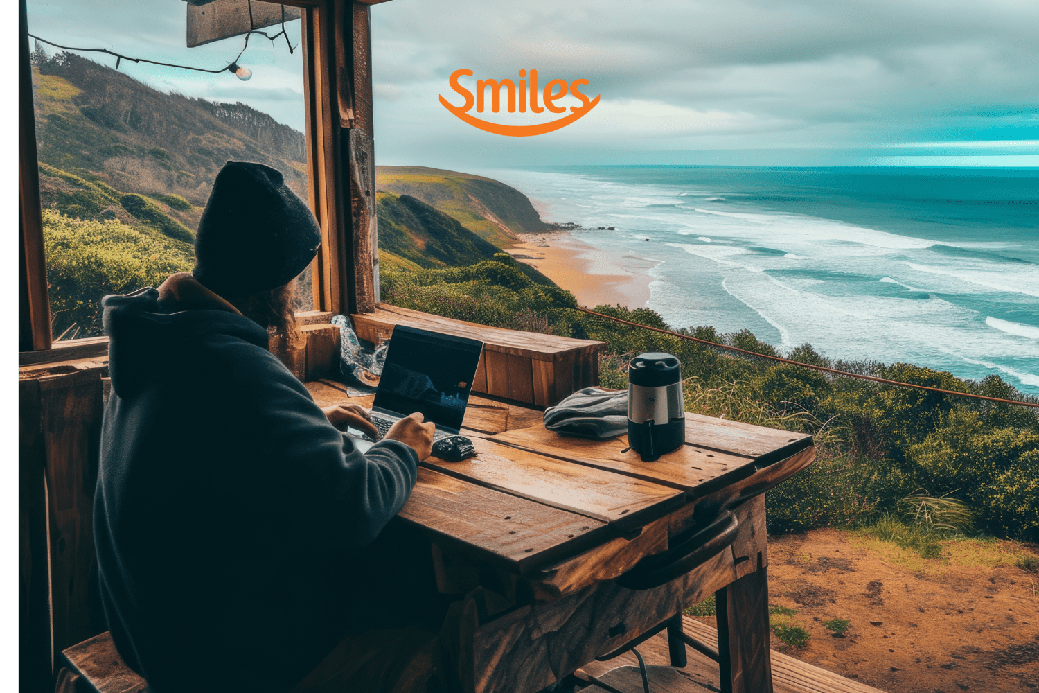"Uma pessoa trabalha em seu laptop em uma cabana de madeira com vista para uma paisagem costeira deslumbrante, onde o mar azul encontra a praia dourada. O clima parece frio e nublado, com uma garrafa térmica sobre a mesa, criando uma atmosfera aconchegante. Acima, o logotipo da Smiles sugere uma conexão com viagens e experiências ao ar livre."