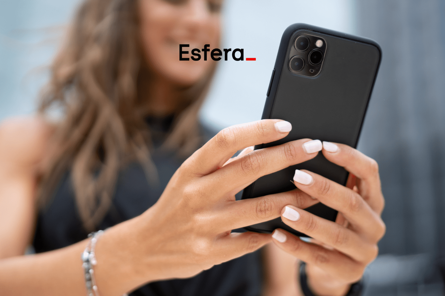 Uma mulher segura um celular preto com as duas mãos, exibindo unhas bem cuidadas e pintadas de branco. O logotipo da Esfera está centralizado na parte superior da imagem, sugerindo uma conexão entre a marca e o uso de tecnologia ou experiências digitais.