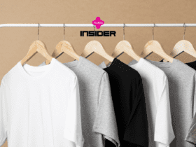 Várias camisetas básicas nas cores branca, cinza e preta estão penduradas em cabides de madeira em uma arara. Acima delas, os logotipos da Livelo e da Insider indicam uma colaboração, sugerindo produtos de moda simples e funcionais, com potencial acúmulo de pontos ou benefícios para os clientes dessas marcas.