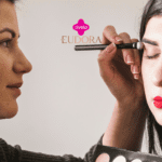 A imagem mostra uma cena de maquiagem, onde uma mulher está aplicando sombra nos olhos de outra mulher, que está com os olhos fechados e usando um batom vermelho intenso. No centro superior da imagem, estão os logotipos da Livelo e da Eudora, sugerindo uma parceria entre essas marcas.