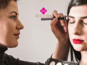 A imagem mostra uma cena de maquiagem, onde uma mulher está aplicando sombra nos olhos de outra mulher, que está com os olhos fechados e usando um batom vermelho intenso. No centro superior da imagem, estão os logotipos da Livelo e da Eudora, sugerindo uma parceria entre essas marcas.