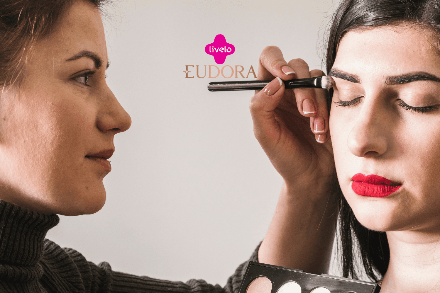 A imagem mostra uma cena de maquiagem, onde uma mulher está aplicando sombra nos olhos de outra mulher, que está com os olhos fechados e usando um batom vermelho intenso. No centro superior da imagem, estão os logotipos da Livelo e da Eudora, sugerindo uma parceria entre essas marcas.