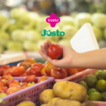 A imagem mostra uma mão segurando um tomate em um ambiente que parece ser uma feira ou mercado, com uma grande variedade de frutas e legumes ao fundo. No centro superior da imagem, estão os logotipos da Livelo e Justo, indicando uma possível colaboração ou promoção entre essas marcas.