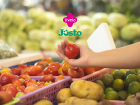 A imagem mostra uma mão segurando um tomate em um ambiente que parece ser uma feira ou mercado, com uma grande variedade de frutas e legumes ao fundo. No centro superior da imagem, estão os logotipos da Livelo e Justo, indicando uma possível colaboração ou promoção entre essas marcas.