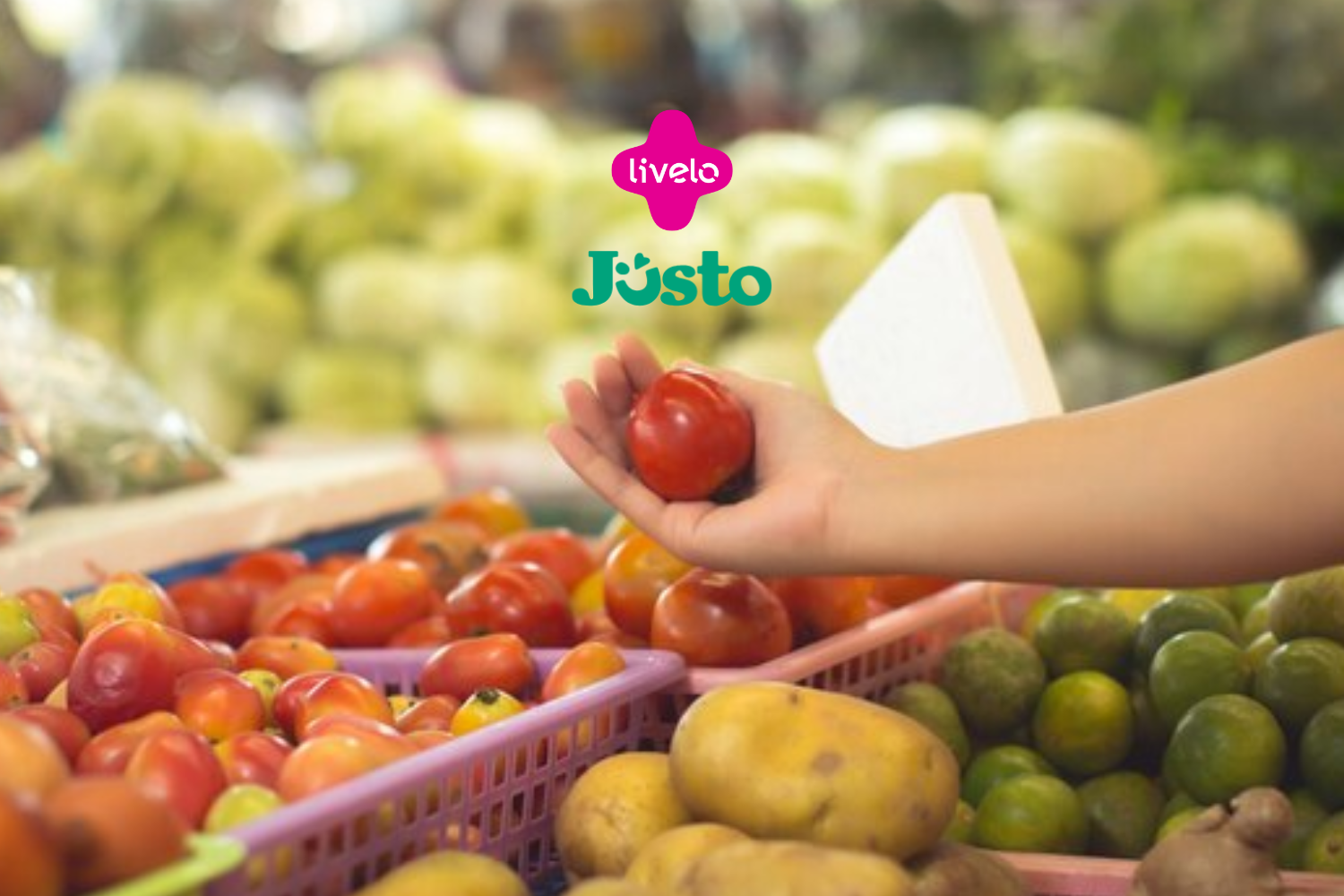 A imagem mostra uma mão segurando um tomate em um ambiente que parece ser uma feira ou mercado, com uma grande variedade de frutas e legumes ao fundo. No centro superior da imagem, estão os logotipos da Livelo e Justo, indicando uma possível colaboração ou promoção entre essas marcas.