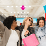 A imagem mostra três mulheres felizes e empolgadas segurando sacolas de compras em um ambiente que parece ser um shopping. As três estão sorrindo e levantando as sacolas, transmitindo uma sensação de alegria e satisfação. No centro superior da imagem, os logotipos da Livelo e da Dudalina estão presentes, sugerindo uma parceria ou promoção entre essas duas marcas.