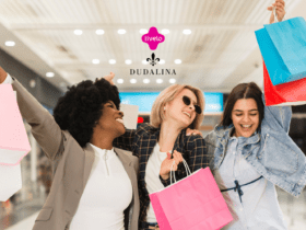 A imagem mostra três mulheres felizes e empolgadas segurando sacolas de compras em um ambiente que parece ser um shopping. As três estão sorrindo e levantando as sacolas, transmitindo uma sensação de alegria e satisfação. No centro superior da imagem, os logotipos da Livelo e da Dudalina estão presentes, sugerindo uma parceria ou promoção entre essas duas marcas.
