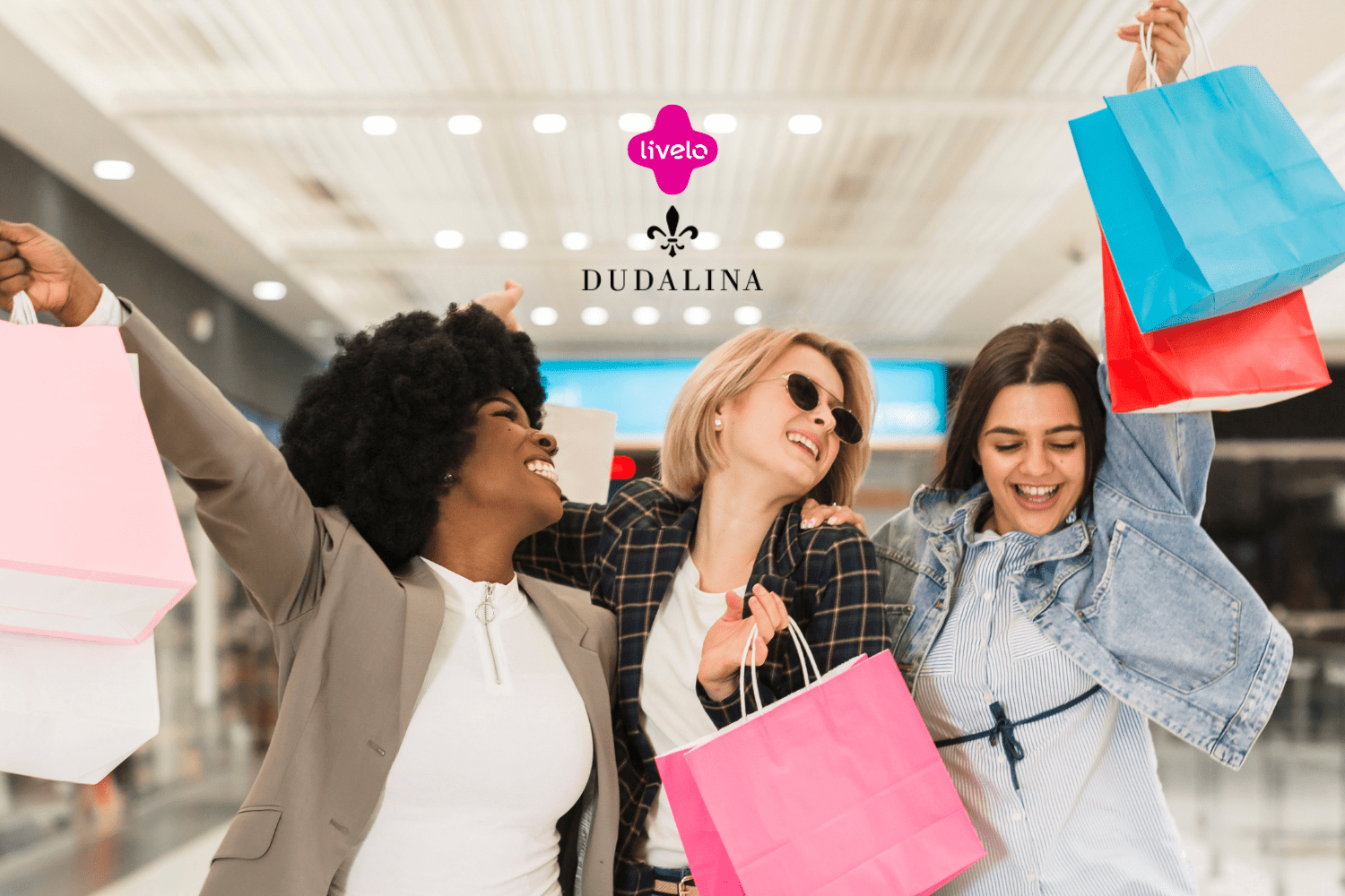 A imagem mostra três mulheres felizes e empolgadas segurando sacolas de compras em um ambiente que parece ser um shopping. As três estão sorrindo e levantando as sacolas, transmitindo uma sensação de alegria e satisfação. No centro superior da imagem, os logotipos da Livelo e da Dudalina estão presentes, sugerindo uma parceria ou promoção entre essas duas marcas.