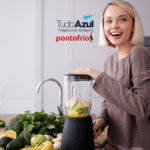 A imagem mostra uma mulher sorridente preparando algo em um liquidificador em uma cozinha moderna. Ao fundo, podemos ver frutas e vegetais frescos, sugerindo que ela está fazendo um suco ou smoothie saudável. No topo da imagem, estão os logotipos do "TudoAzul Programa de Vantagens" e "pontofrio", indicando uma possível promoção ou parceria entre essas marcas.