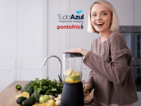 A imagem mostra uma mulher sorridente preparando algo em um liquidificador em uma cozinha moderna. Ao fundo, podemos ver frutas e vegetais frescos, sugerindo que ela está fazendo um suco ou smoothie saudável. No topo da imagem, estão os logotipos do "TudoAzul Programa de Vantagens" e "pontofrio", indicando uma possível promoção ou parceria entre essas marcas.