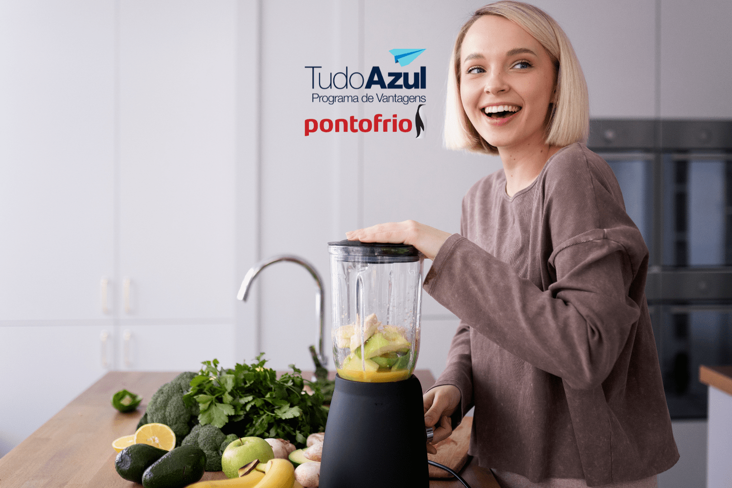 A imagem mostra uma mulher sorridente preparando algo em um liquidificador em uma cozinha moderna. Ao fundo, podemos ver frutas e vegetais frescos, sugerindo que ela está fazendo um suco ou smoothie saudável. No topo da imagem, estão os logotipos do "TudoAzul Programa de Vantagens" e "pontofrio", indicando uma possível promoção ou parceria entre essas marcas.
