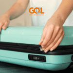 Pessoa fechando uma mala de viagem azul-clara com um cadeado embutido. Ao fundo, o logotipo da GOL Linhas Aéreas, destacando o tema de transporte aéreo e bagagem. despacho de bagagem