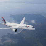 Avião da LATAM sobrevoando uma região costeira com montanhas e mar ao fundo, destacando o design moderno da aeronave e sua marca icônica.