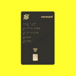 Imagem de um cartão BB Voz Ourocard preto com detalhes em dourado, incluindo o logotipo do Banco do Brasil e informações em braille. Fundo amarelo vibrante.