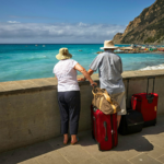Casal de idosos com chapéus observando o mar azul de um mirante, com malas de viagem ao lado, em um destino litorâneo cercado por montanhas com o Programa Voa Brasil.