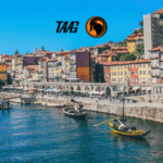 Vista do Cais da Ribeira, na cidade do Porto, Portugal, com barcos tradicionais no Rio Douro e fachadas coloridas ao fundo, acompanhada do logotipo da TAAG Angola Airlines no céu azul.