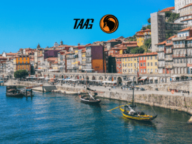 Vista do Cais da Ribeira, na cidade do Porto, Portugal, com barcos tradicionais no Rio Douro e fachadas coloridas ao fundo, acompanhada do logotipo da TAAG Angola Airlines no céu azul.