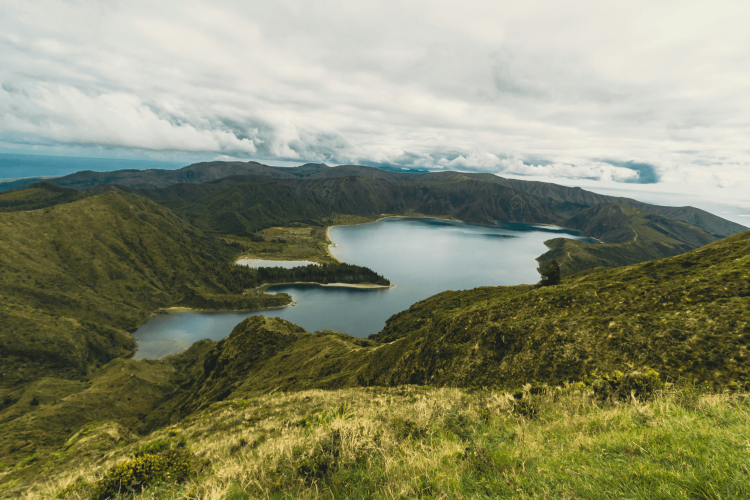 Vista panorâmica da Lagoa do Fogo cercada por montanhas verdes e céu nublado, um dos cenários mais emblemáticos de onde fica a Ilha de São Miguel, em Portugal.